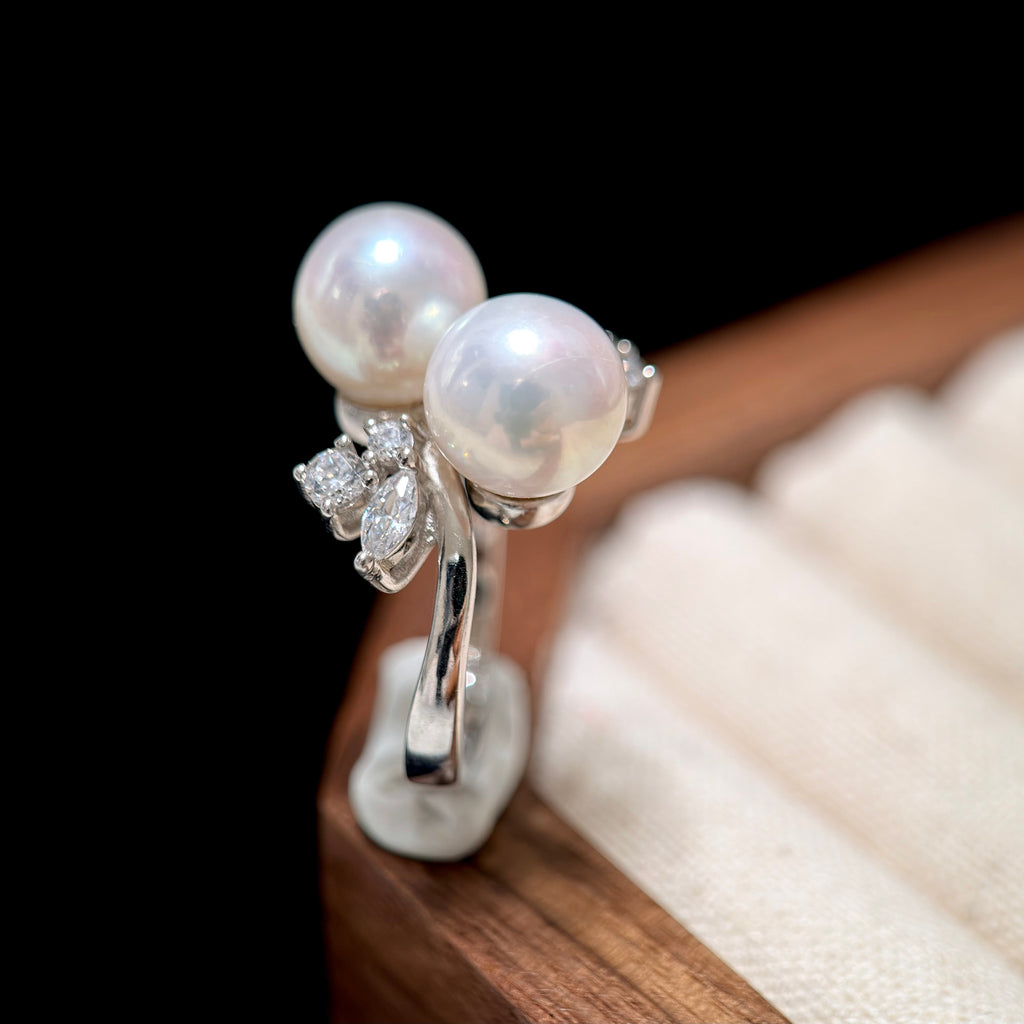 A silver ring featuring two large white pearls and a small cluster of clear stones, displayed on a stand against a dark background with a blurred jewelry box.