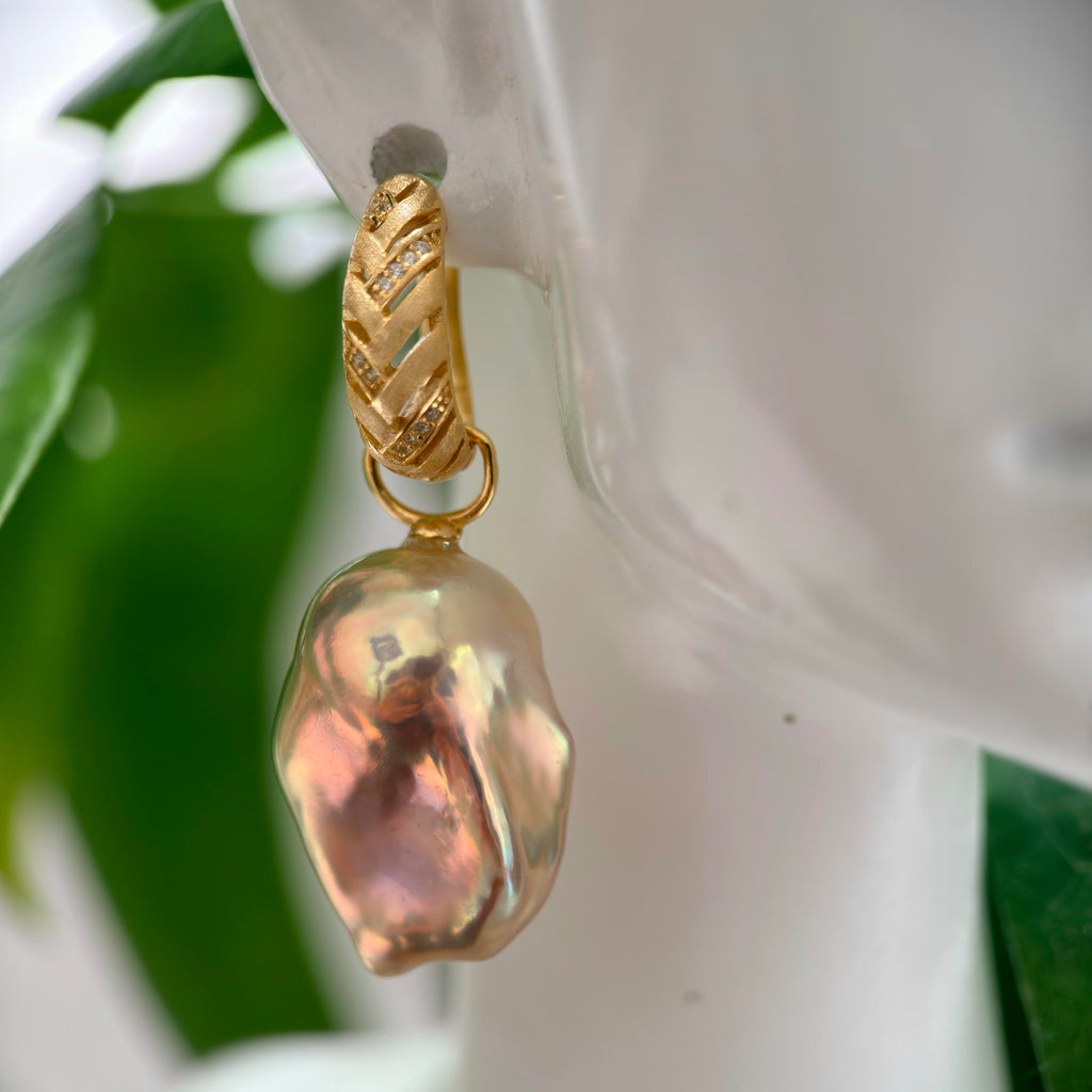 A gold hoop earring with a baroque pearl drop, displayed on a white mannequin head with green leaves in the background.