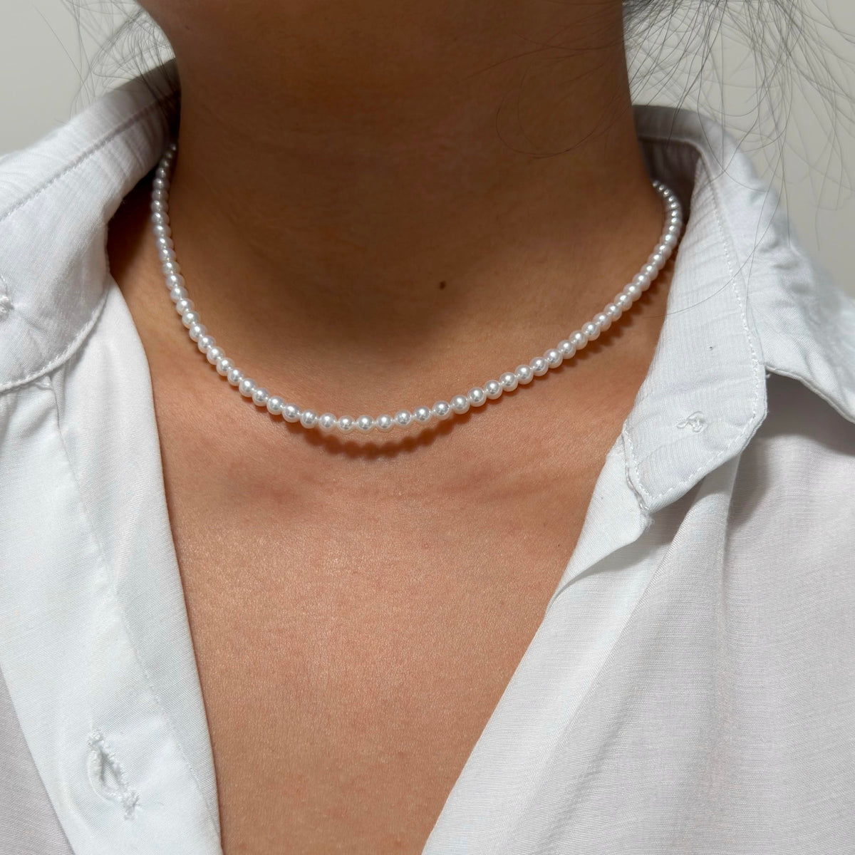 A close-up of a person wearing a delicate bright white pearl choker necklace with a white collared shirt.