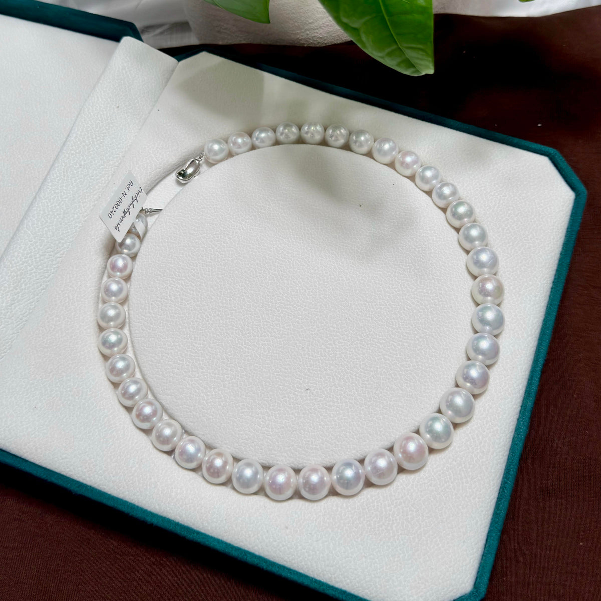 A freshwater pearl necklace arranged in a circle inside an open white jewelry box with a dark teal border. A small paper tag is attached near the clasp, and a green leaf is visible at the top edge of the frame.