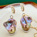 Gold huggie hoop earrings and matching pendant necklace featuring large baroque pearl charms with iridescent purple, green, and gold tones, displayed on a jewelry stand.