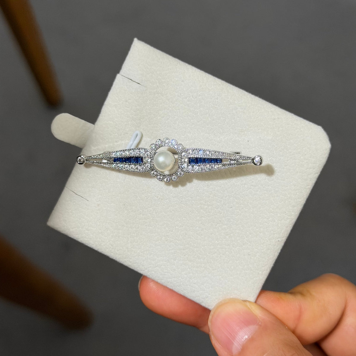 A silver-toned Art Deco style brooch featuring a central white pearl and blue sapphires, set with small clear crystals, displayed on a cream jewelry card held by a hand.