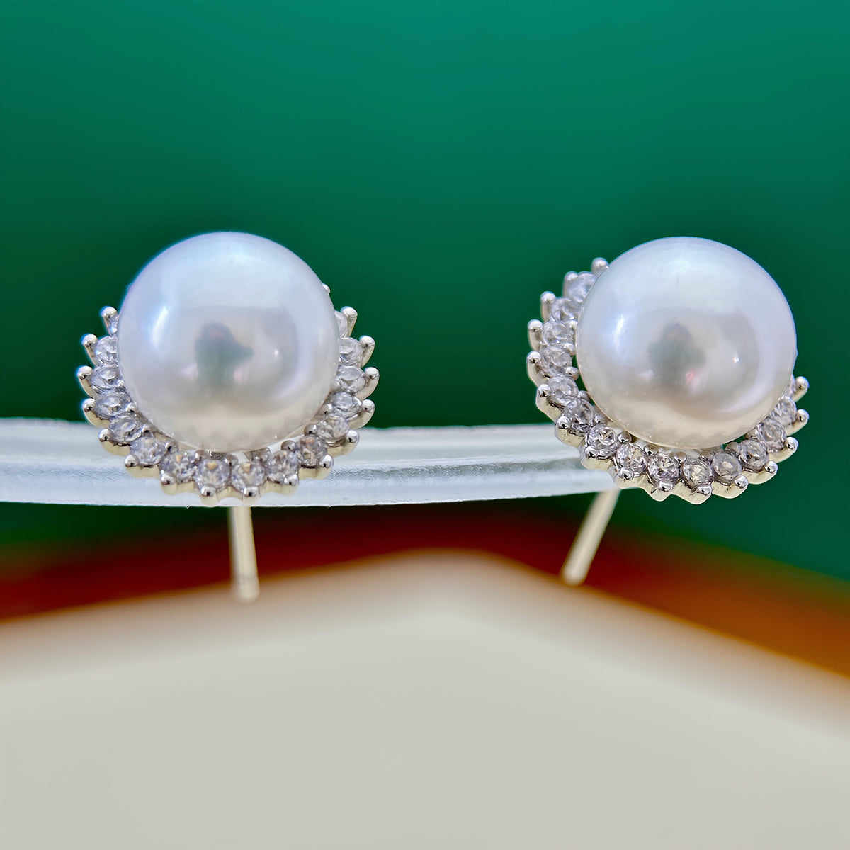A pair of pearl stud earrings with silver-toned halo settings, displayed on a clear stand against a green and beige background.