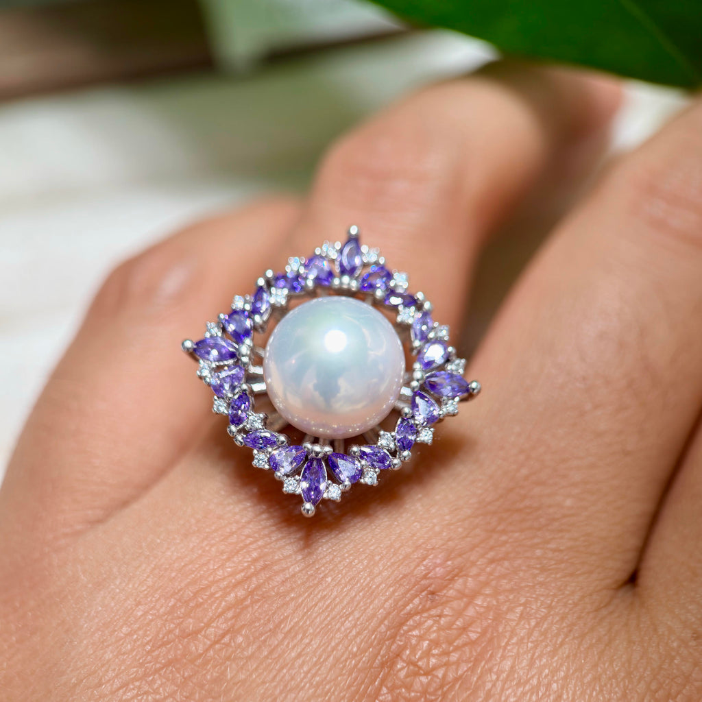 A close-up of a ring worn on a finger, featuring a large round white pearl surrounded by a halo of small purple and clear stones in a silver-toned setting.