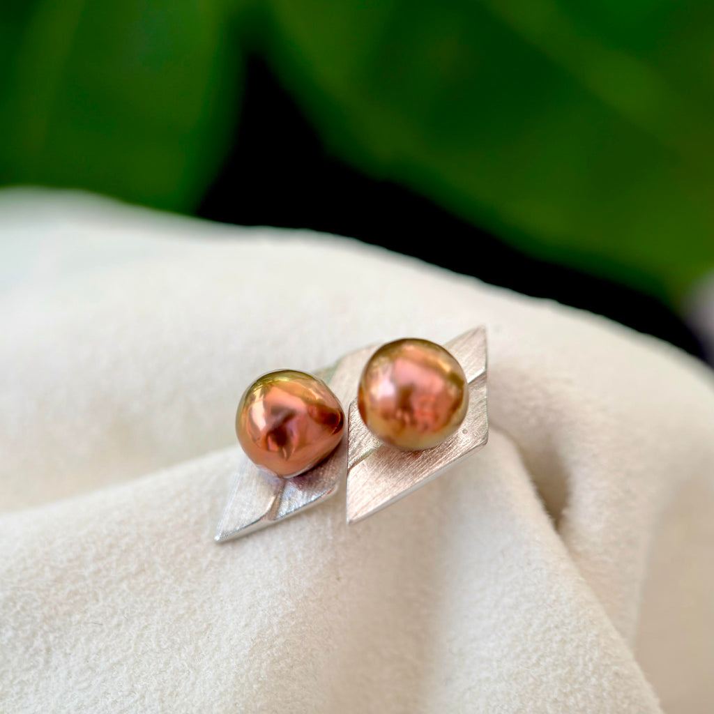 A pair of silver leaf-shaped stud earrings with iridescent coppery-brown freshwater pearls, displayed on a soft white fabric with a blurred green background.