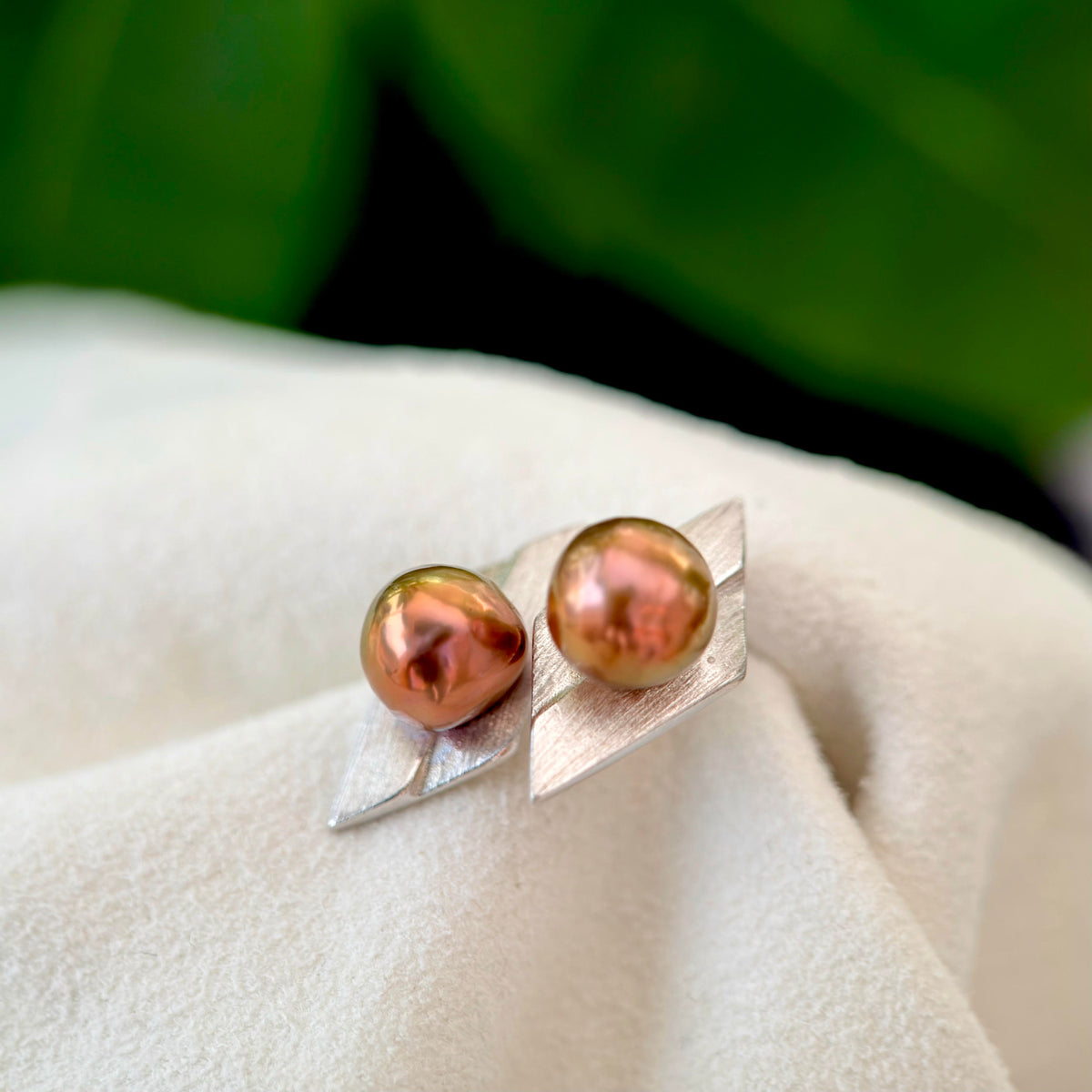 A pair of silver leaf-shaped stud earrings with iridescent coppery-brown freshwater pearls, displayed on a soft white fabric with a blurred green background.