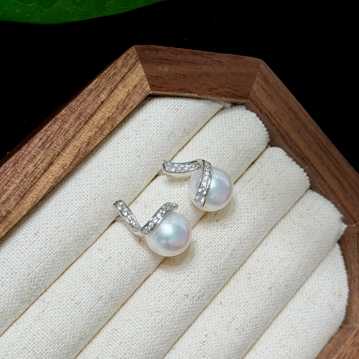 A pair of silver hoop earrings with white freshwater pearls and small clear stones, displayed on a cream fabric-lined wooden jewelry box.