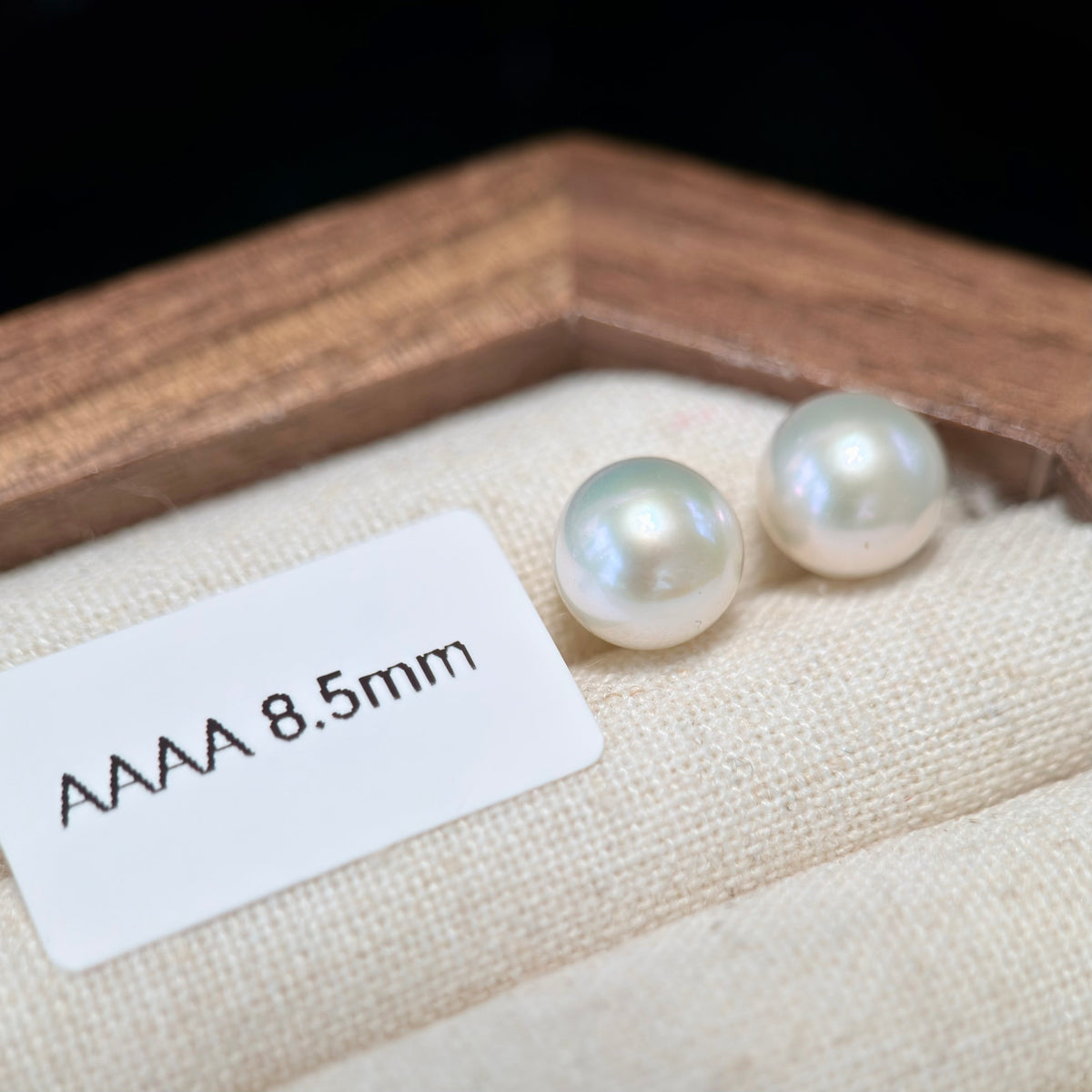 Two round AAAA grade 8.5mm freshwater pearls displayed on a beige fabric surface next to a white size label, with a wooden box frame in the background.