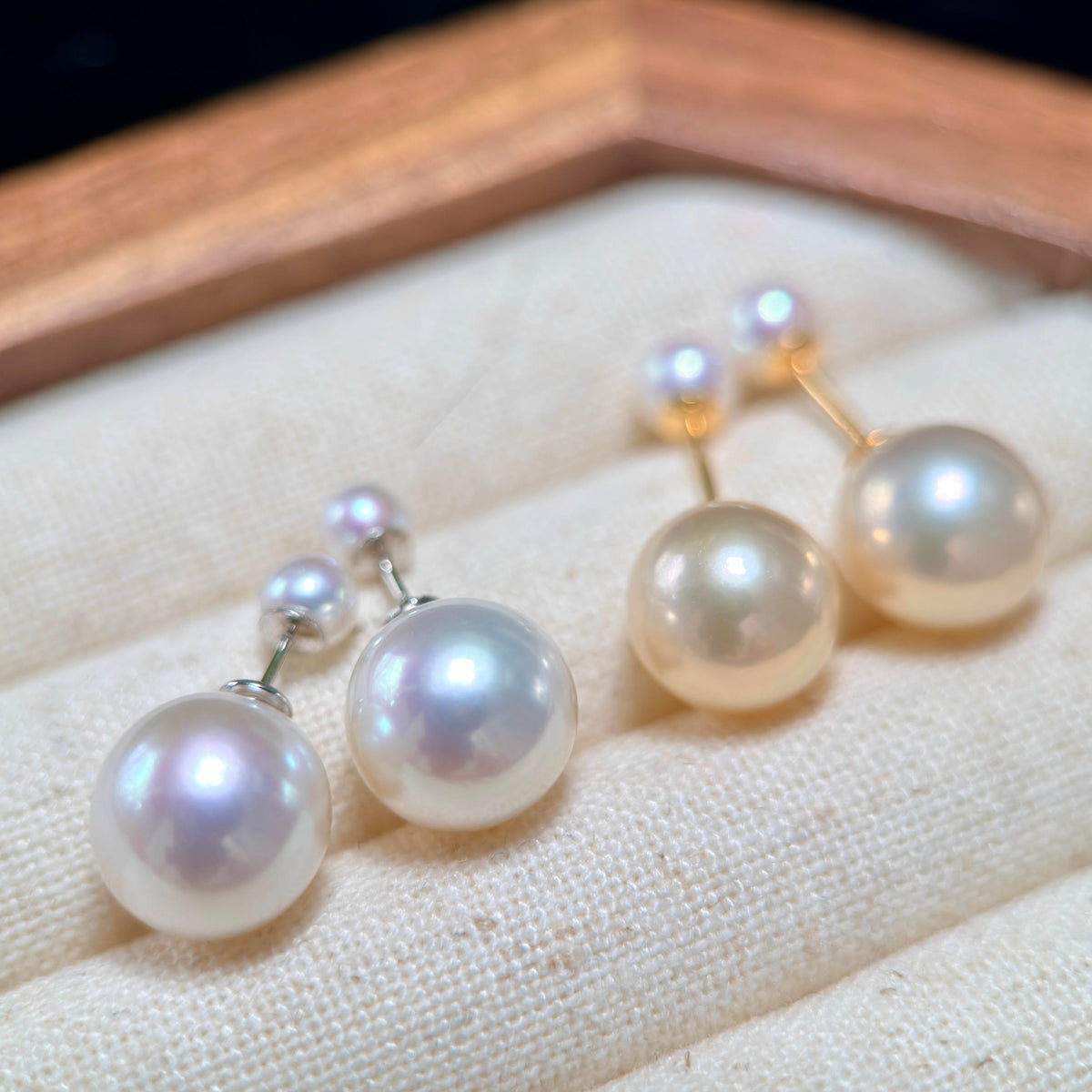 A pair of pearl stud earrings with small pearl accents and gold-tone posts, displayed on a cream fabric surface inside a wooden box.