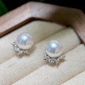 A pair of pearl stud earrings with silver leaf-shaped settings accented by small clear crystals, displayed on a cream fabric jewelry cushion inside a wooden box.