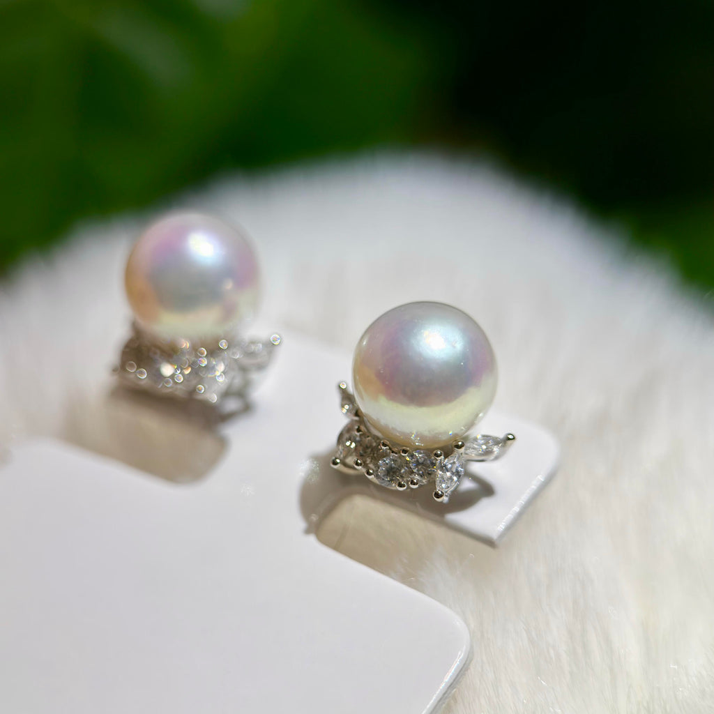 A pair of pearl stud earrings with silver floral settings, displayed on a white card over a soft white faux fur surface with a blurred green background.