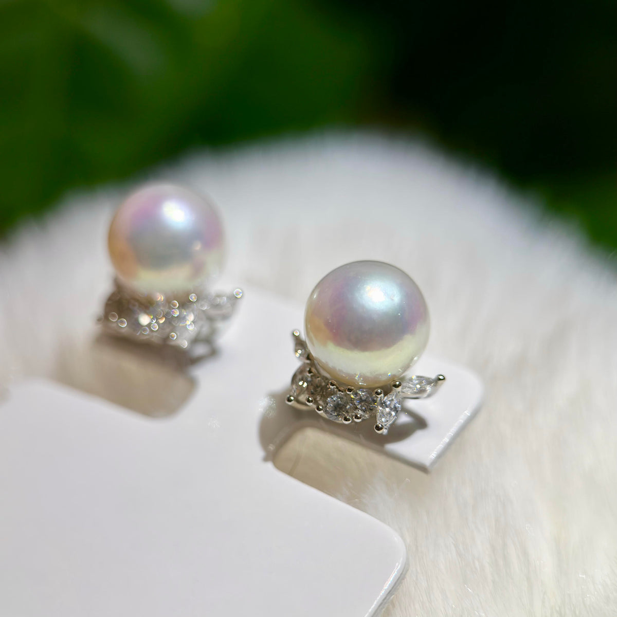 A pair of pearl stud earrings with silver floral settings, displayed on a white card over a soft white faux fur surface with a blurred green background.