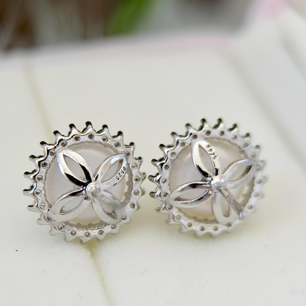 A pair of silver butterfly stud earrings with scalloped circular borders, displayed on a light cream surface.