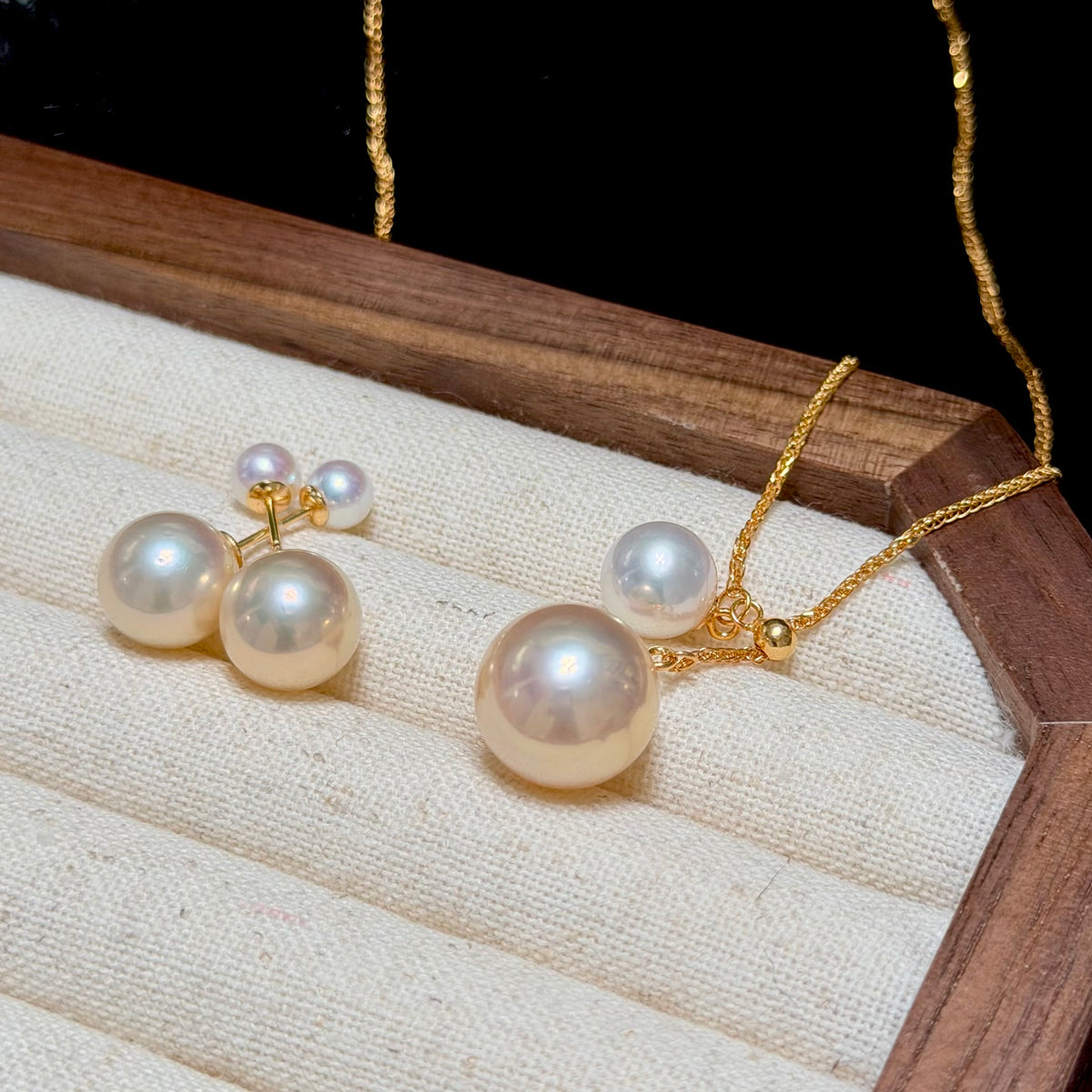 A pearl jewelry set displayed on a cream fabric-lined wooden tray, featuring a gold necklace with a large round pearl pendant and matching pearl stud earrings with gold posts.
