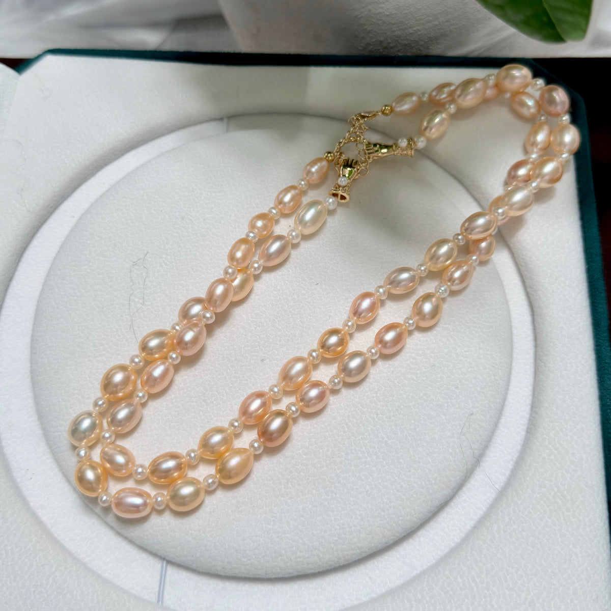 A double-strand pearl necklace with light pinkish-beige pearls and gold-toned clasp, displayed on a white circular jewelry stand.