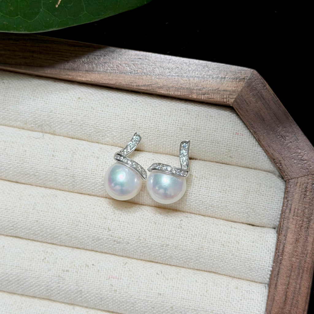 A pair of silver-toned earrings with oval white freshwater pearls and small clear stones, displayed on a cream fabric jewelry tray inside a wooden box.