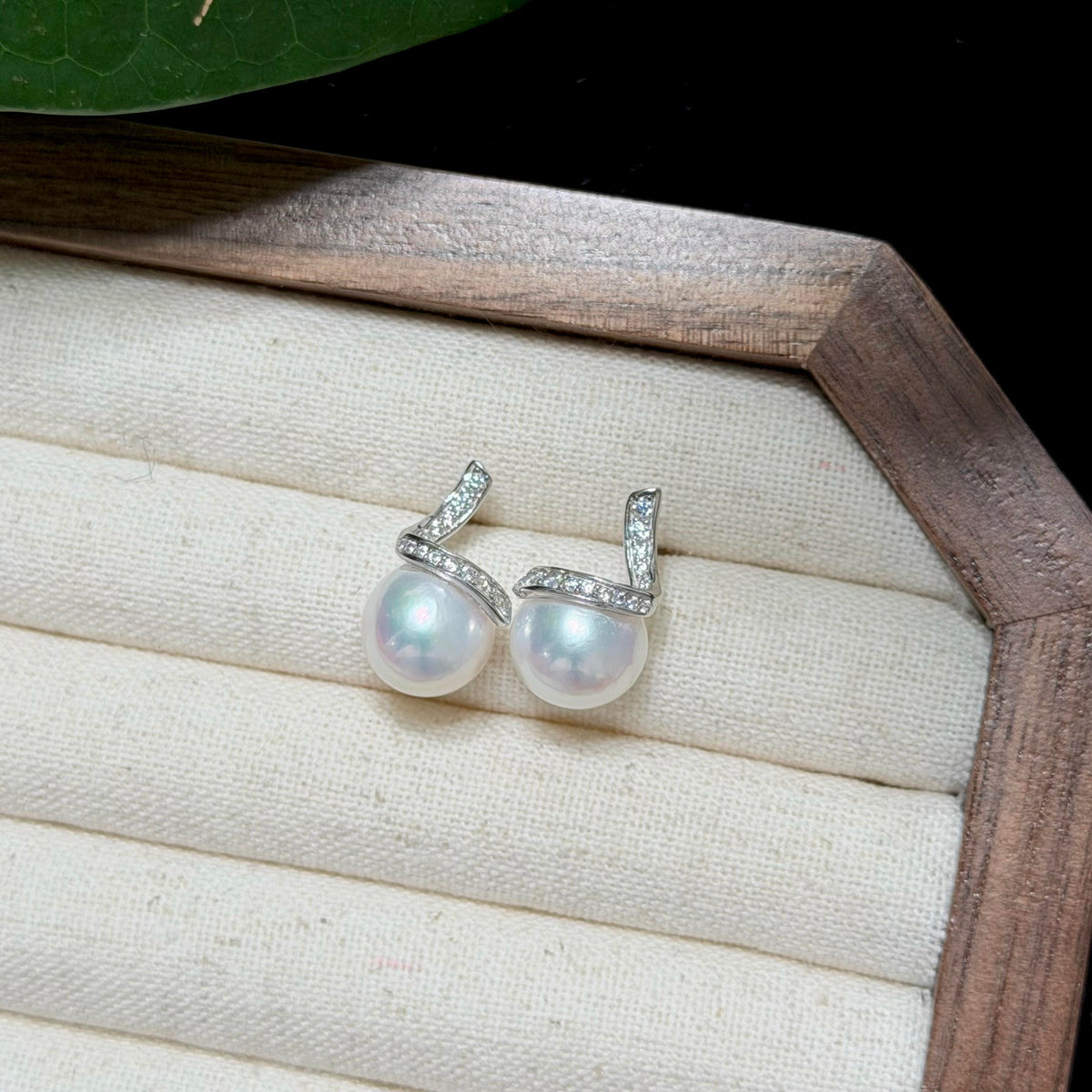 A pair of silver-toned earrings with oval white freshwater pearls and small clear stones, displayed on a cream fabric jewelry tray inside a wooden box.