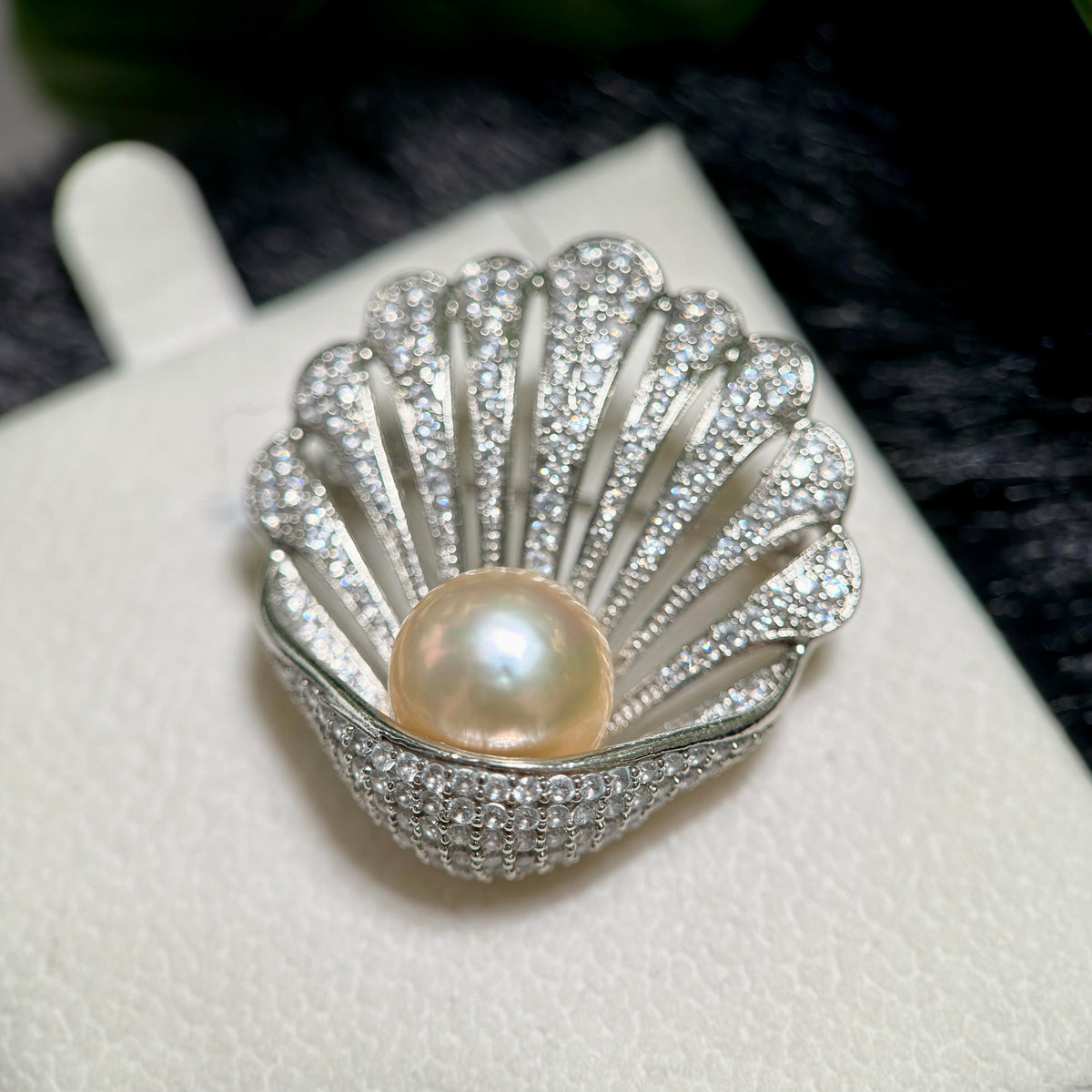 A shell-shaped silver ring brooch or pendant featuring a central freshwater pearl and pavé-set clear stones, displayed on a white jewelry card.