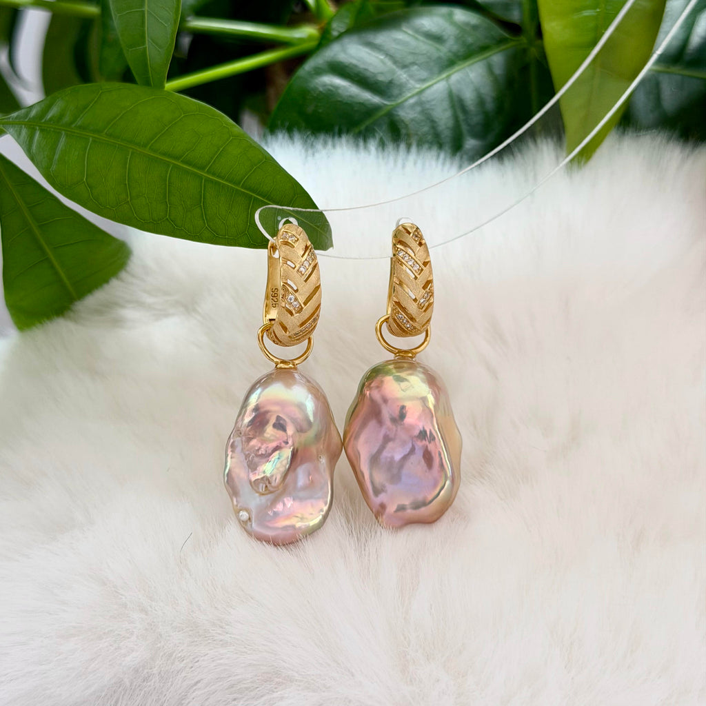 A pair of gold hoop earrings with large, organic-shaped iridescent pink baroque pearls, displayed on a white faux fur surface with green leaves in the background.