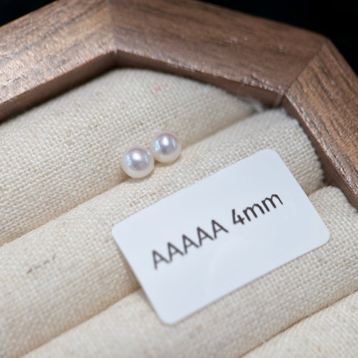 Two small round white pearls displayed on a beige fabric jewelry tray inside a wooden box, with a white size card reading 'AAAAA 4mm'.