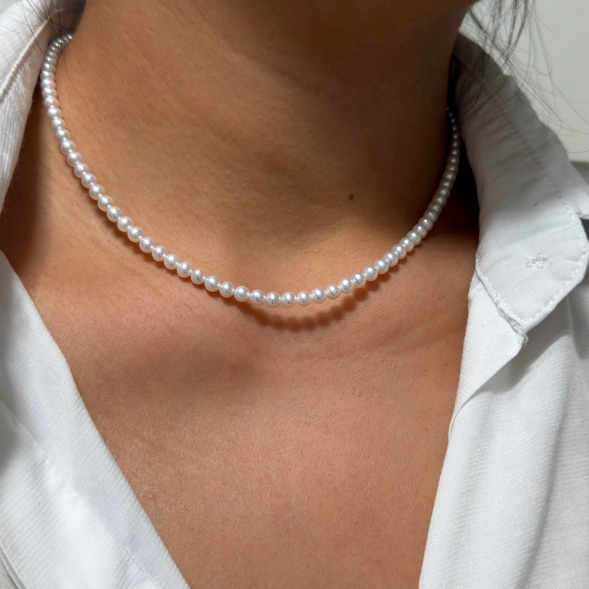 A close-up of a person wearing a classic single-strand white pearl choker necklace with a white collared shirt.