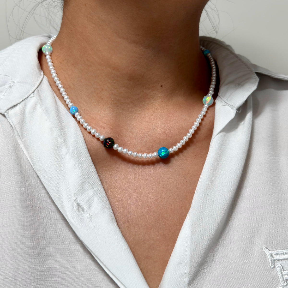 A close-up of a person wearing a beaded necklace over a white collared shirt. The necklace features white pearl-like beads with accent beads in turquoise blue, black, and light green.