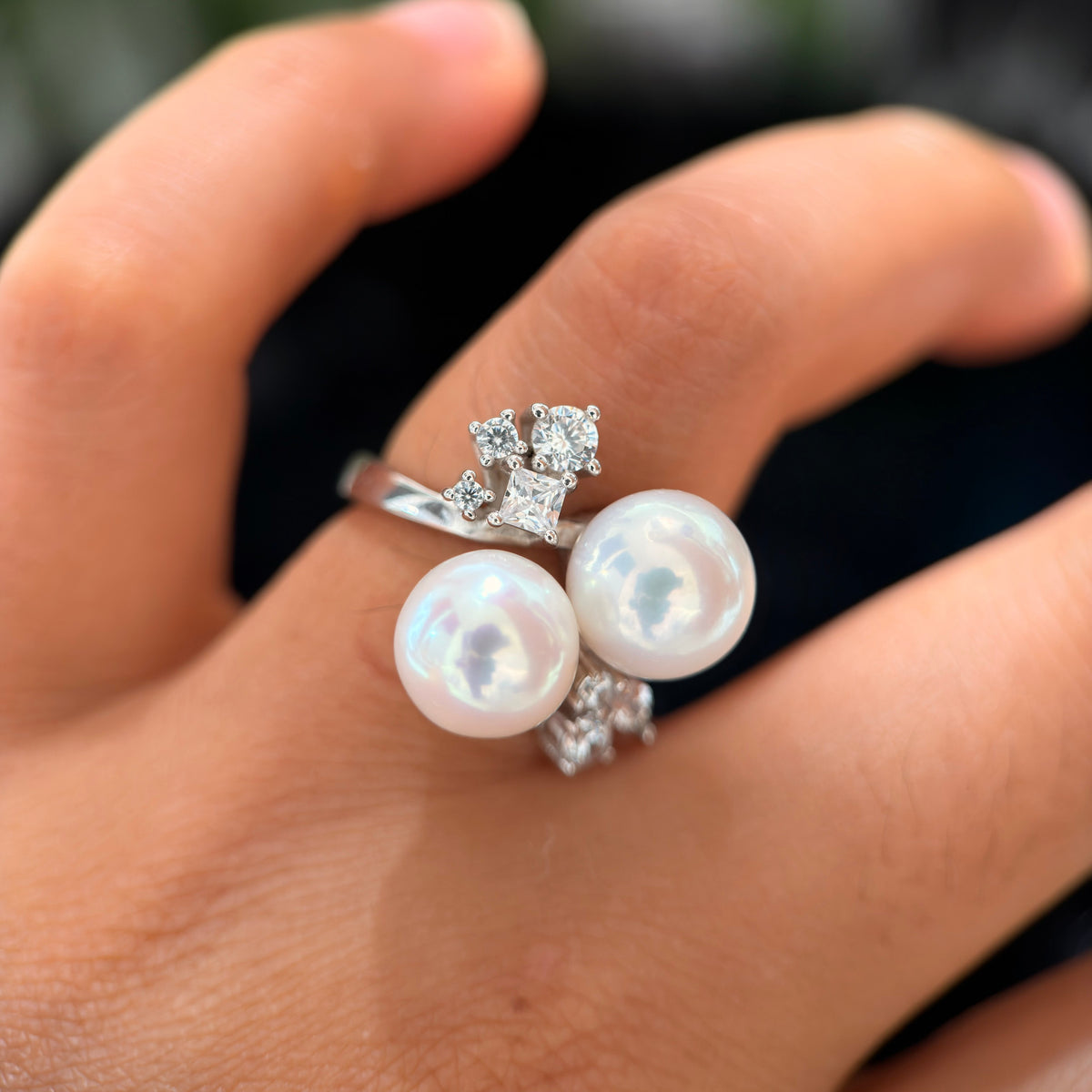A close-up of a hand wearing a silver ring with two large white pearls and small clear gemstones.