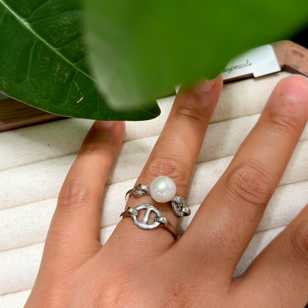 A close-up of a hand wearing three stacked silver rings, including a ring with a white pearl and textured silver bands, resting on a cream-colored ring box with a green leaf in the foreground.