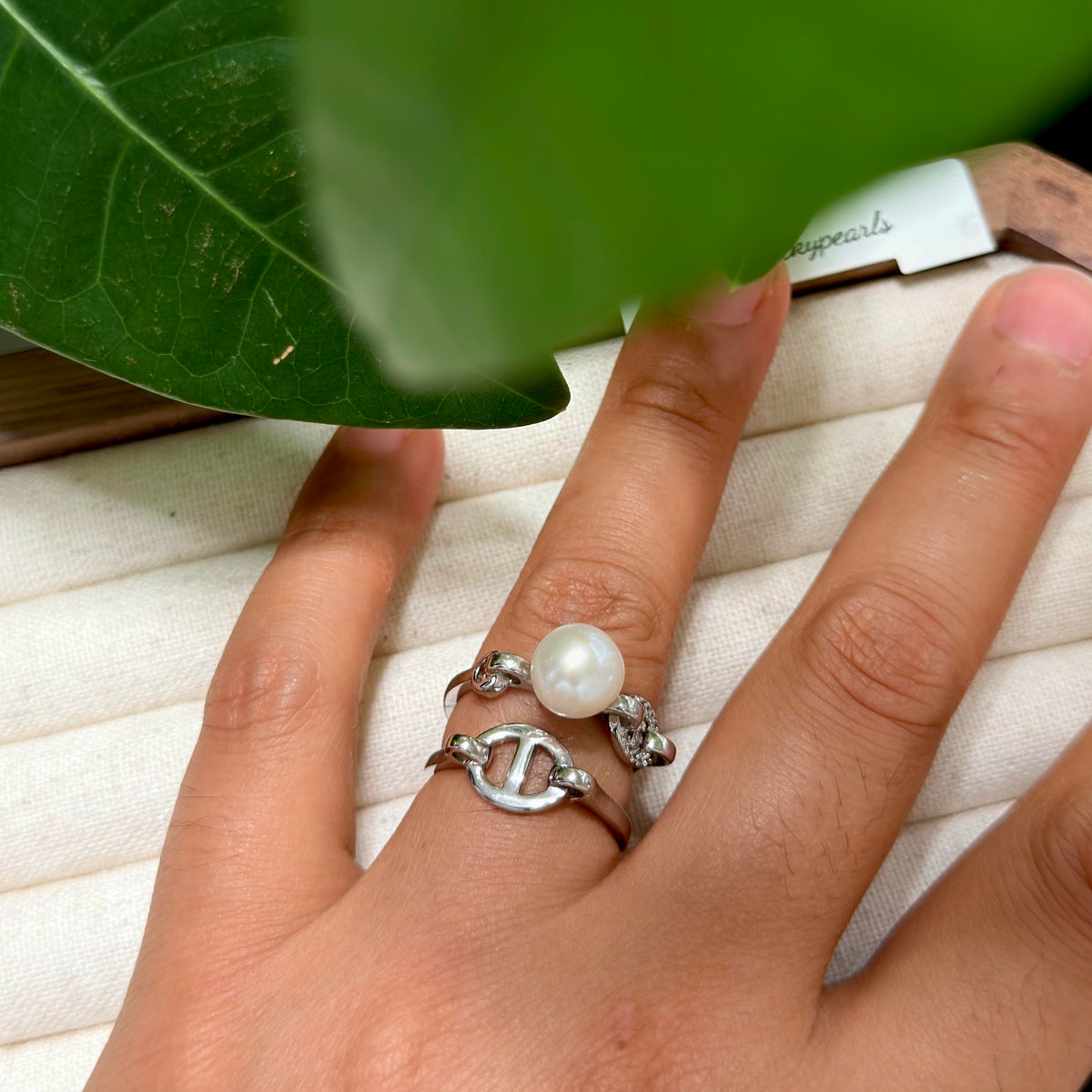 A close-up of a hand wearing three stacked silver rings, including a ring with a white pearl and textured silver bands, resting on a cream-colored ring box with a green leaf in the foreground.
