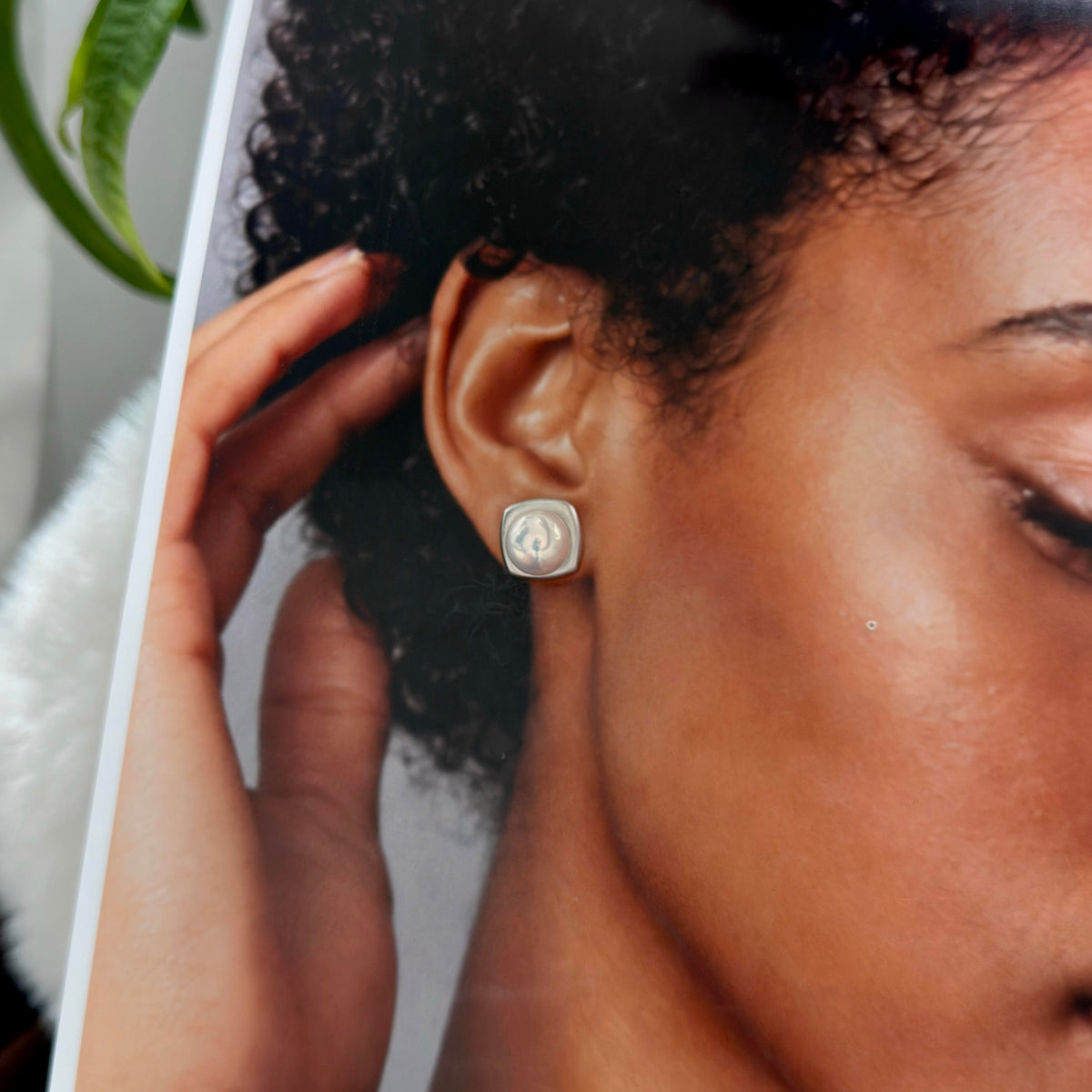 A close-up of a person wearing a small square stud earring with a silver-toned metal border and a white inlay or stone in the center. The photo is cropped tightly on the ear and side of the face, with part of a hand visible adjusting the earring.