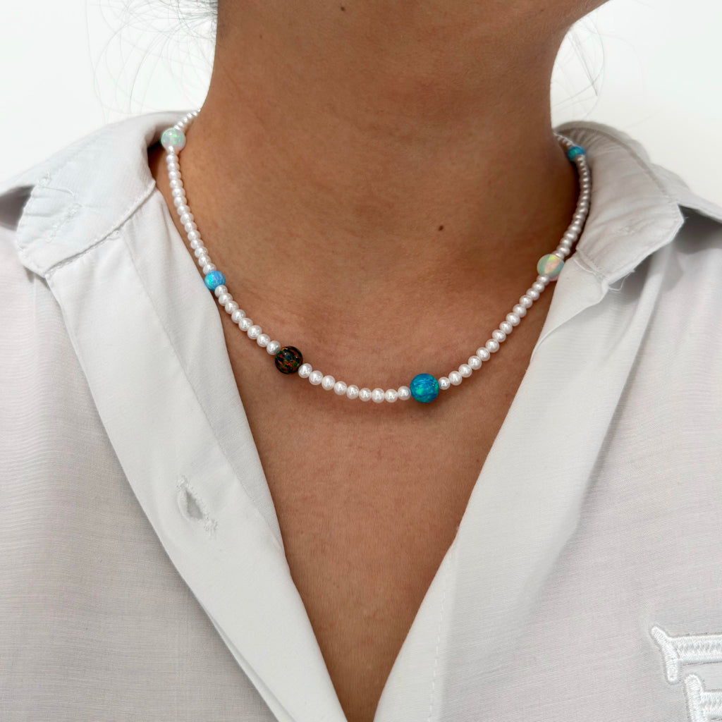 A close-up of a person wearing a delicate necklace made of small white pearls with turquoise blue beads and a few dark accent opal, paired with a white collared shirt.