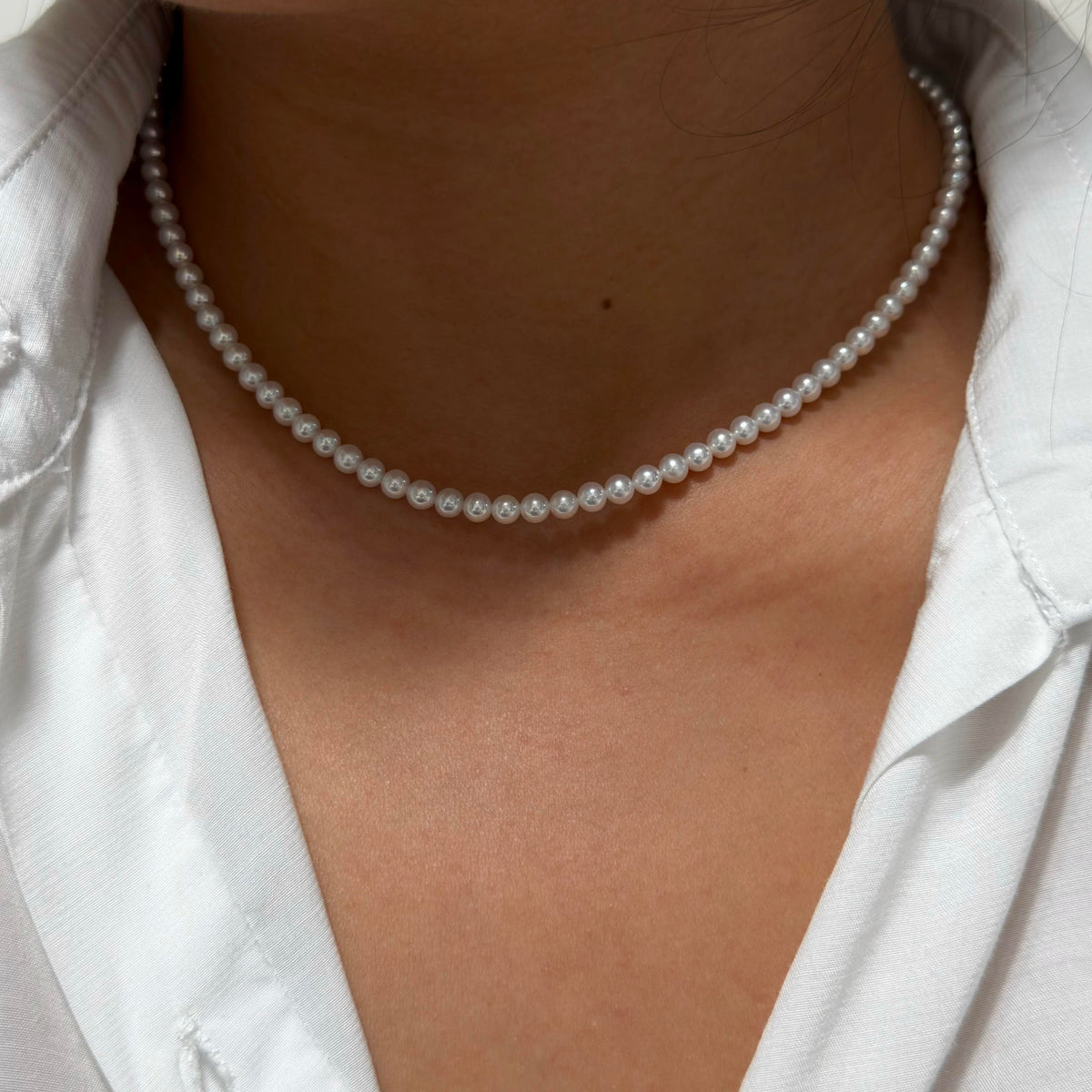 A close-up of a person wearing a delicate single-strand pearl choker necklace with a white button-down shirt.