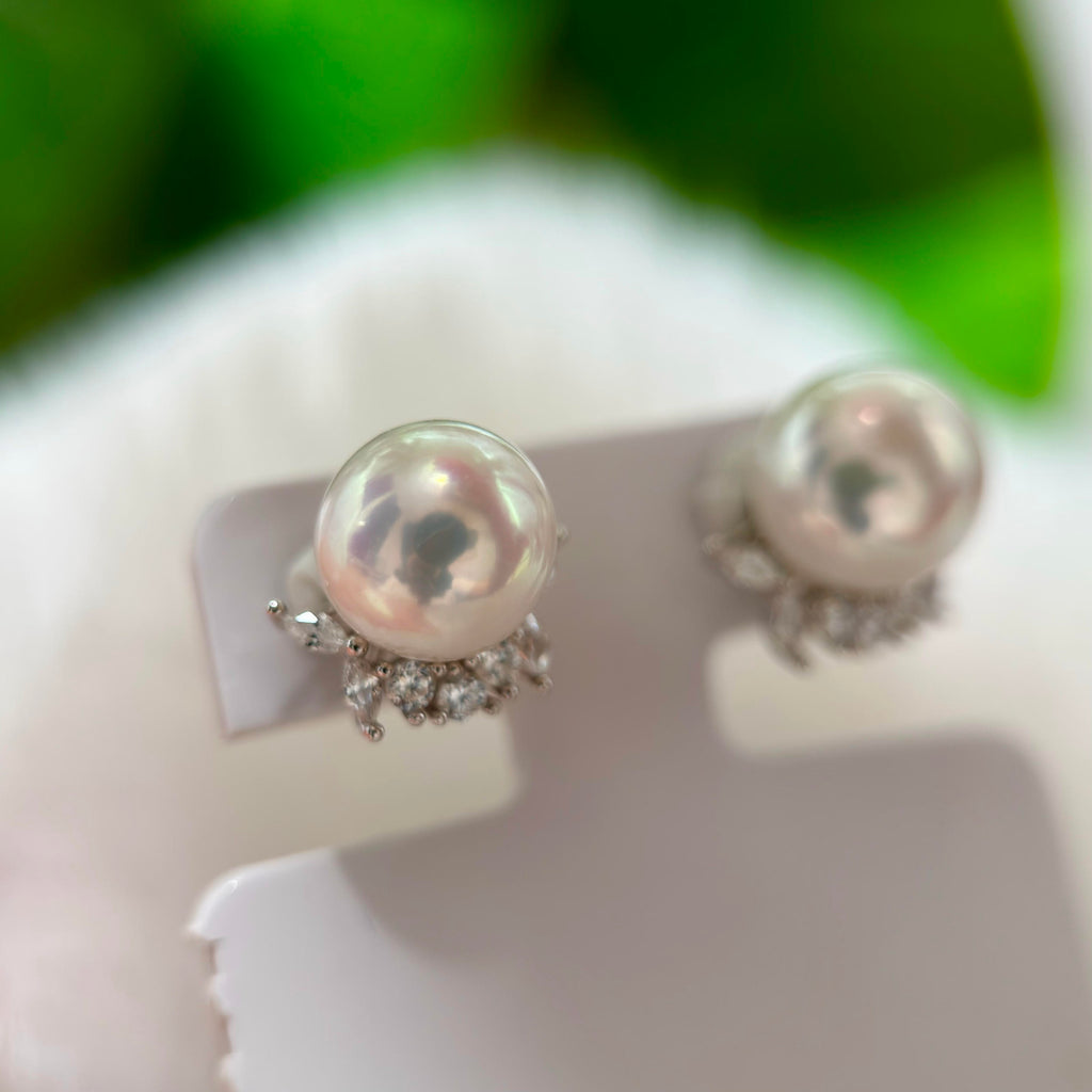A pair of pearl stud earrings with silver-toned floral settings and small clear stones, displayed on a white stand against a soft blurred green and white background.