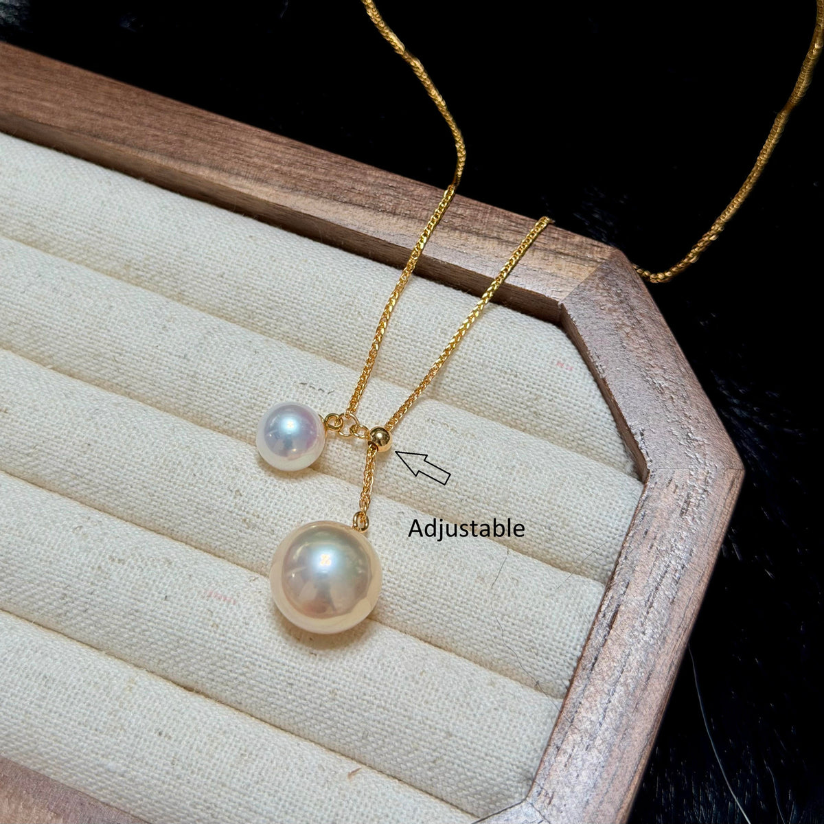 Gold necklace with two pearl-like drops, one larger and one smaller, displayed on a cream jewelry cushion inside a wooden box. A small arrow points to the adjustable clasp area with the text 'Adjustable'.