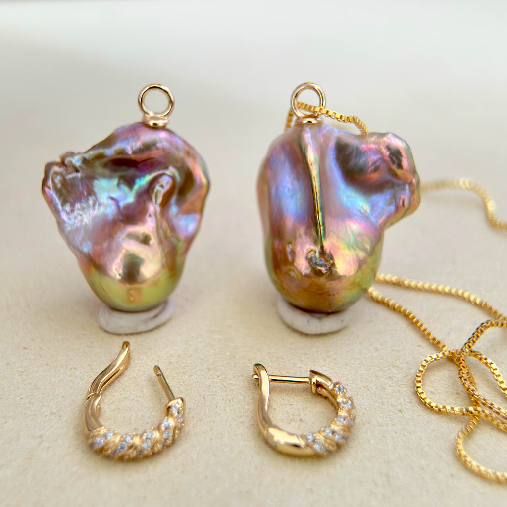 A pair of gold-toned baroque pearl pendants with matching hoop earrings and a gold chain, displayed on a neutral beige surface.