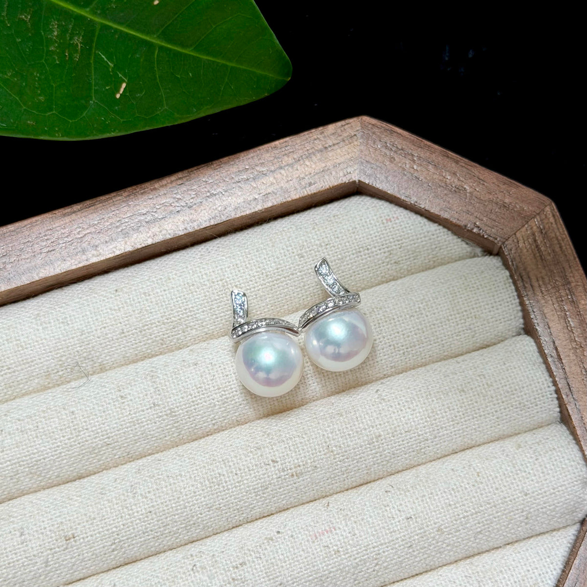 A pair of silver-toned drop earrings with round white pearls and small clear stones, displayed on a cream fabric-lined jewelry box with a wooden frame.