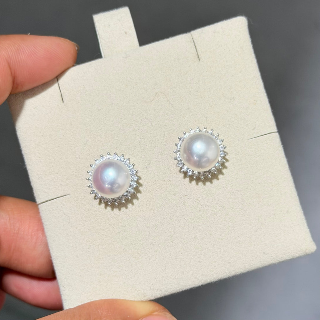 A pair of silver stud earrings with round white pearls surrounded by small clear crystals, displayed on a beige jewelry card held by a hand.