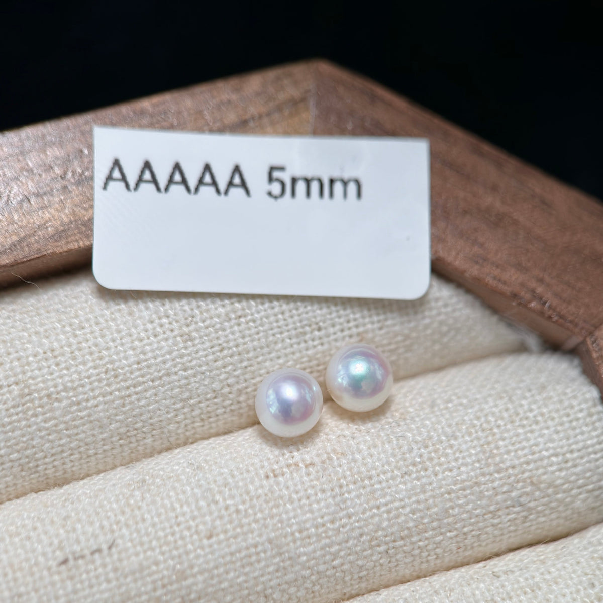 Two round freshwater pearls, labeled AAAAA 5mm, displayed on a beige fabric jewelry cushion inside a wooden box.