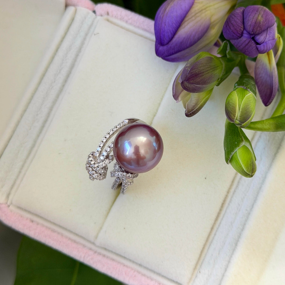 A ring with a large purple pearl and a silver-toned pavé band, displayed on a cream-colored jewelry box with purple flower buds as props.
