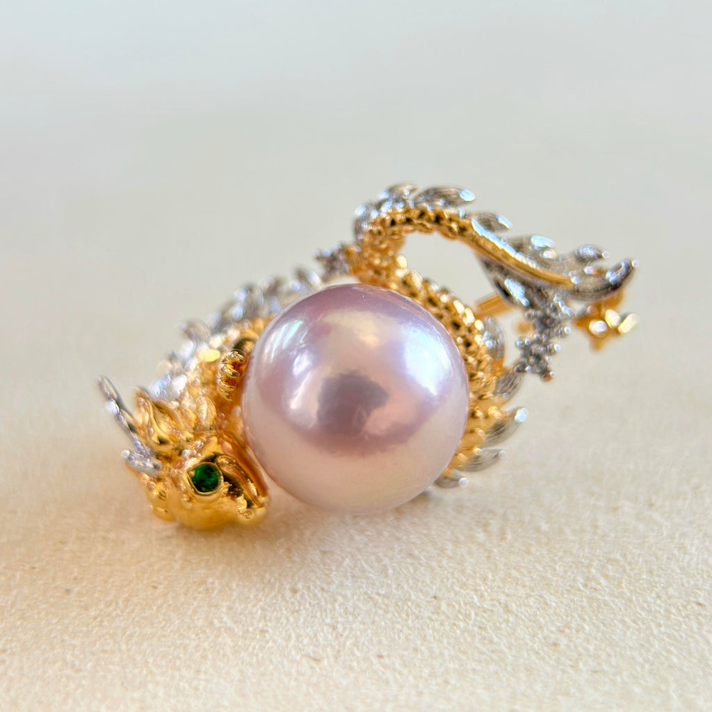 An ornate dragon-themed ring featuring a large lustrous pinkish pearl, gold-toned dragon head and body, silver-toned dragon wings and tail, and a small green gemstone in one eye, set against a soft beige background.
