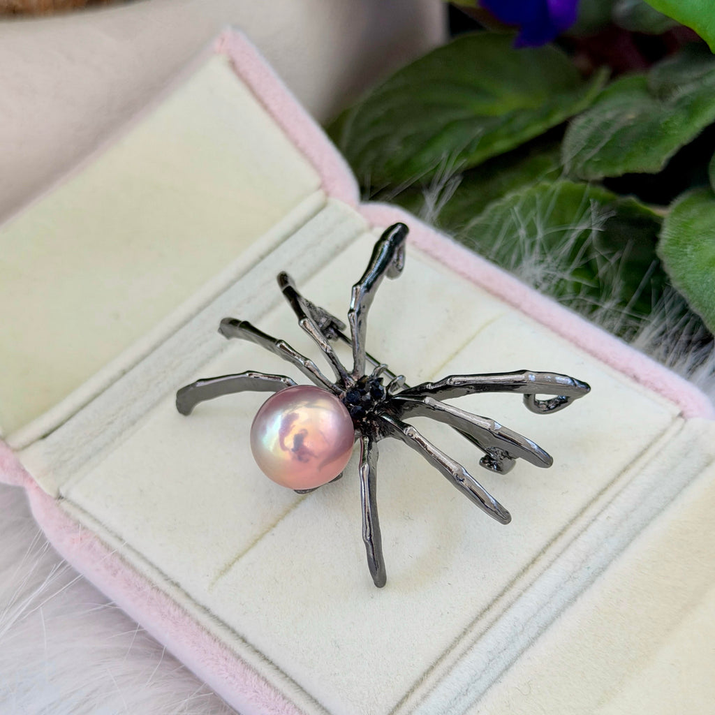 A spider-shaped brooch with a pink pearl and dark metallic legs, displayed on an open pink jewelry box with green leaves in the background.
