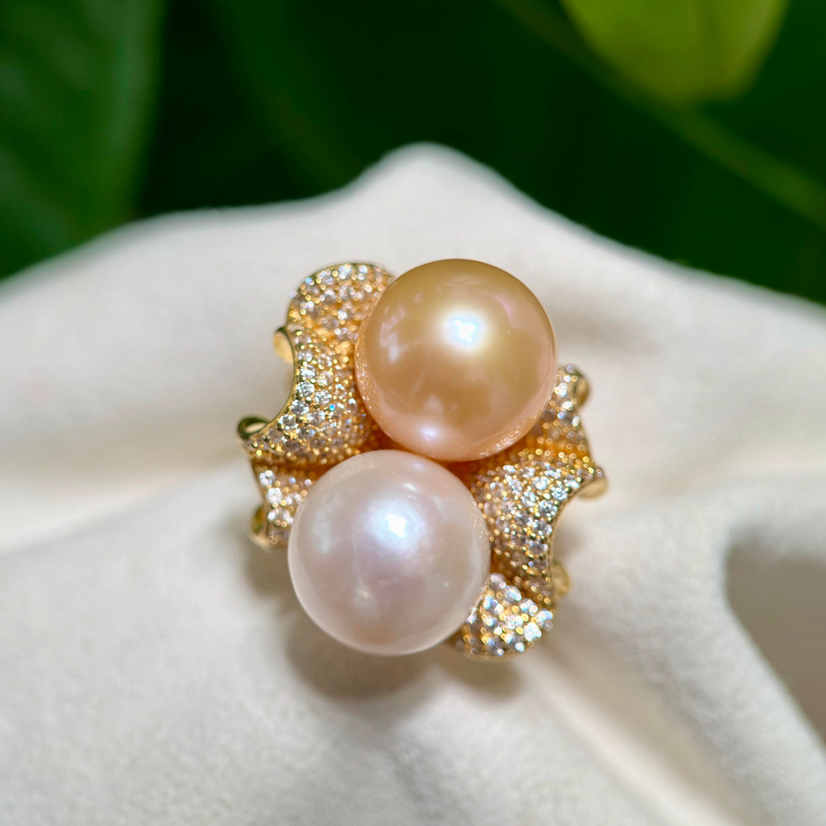 A gold ring featuring two large round freshwater pearls, one pearlescent cream and one soft pink, set in a textured pavé setting on a white fabric surface with a blurred green background.