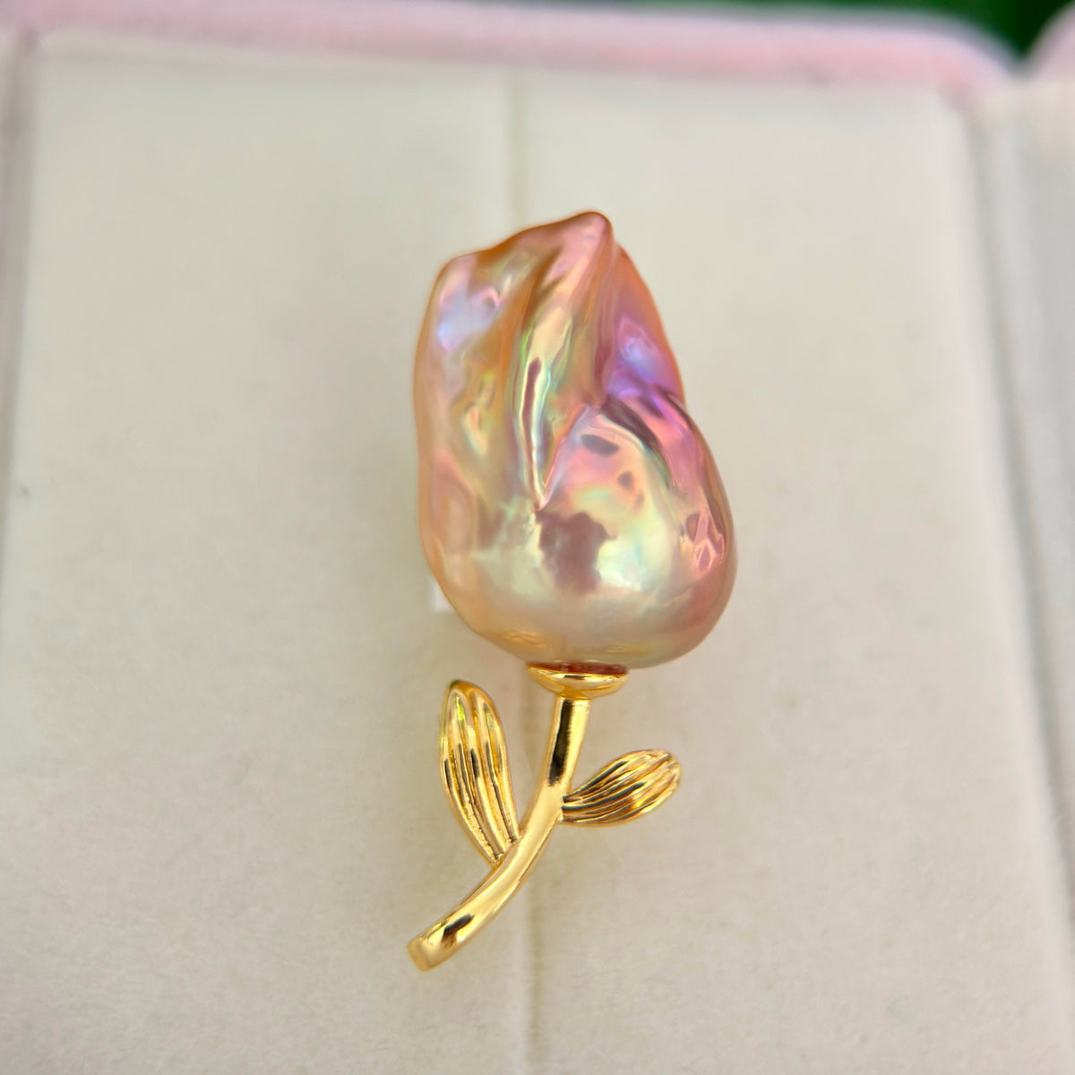 A baroque pearl rose brooch with a gold stem and leaves, displayed on a light cream jewelry box.