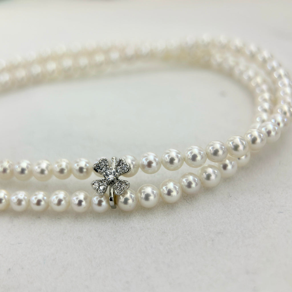 A double-strand pearl necklace with a silver-toned four-leaf clover charm embellished with clear rhinestones, displayed on a light textured surface.