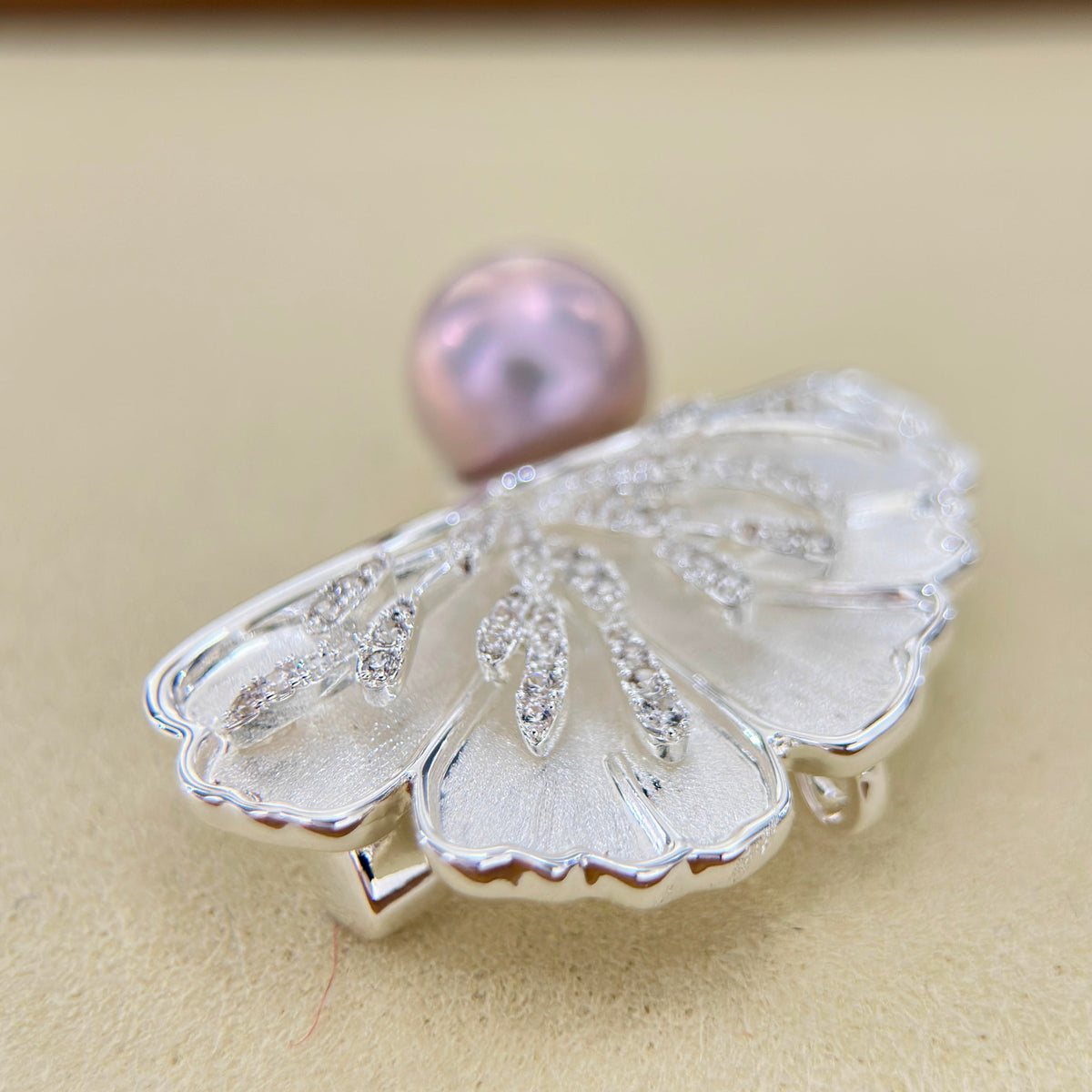 A silver butterfly-shaped brooch featuring a central iridescent pink pearl and small clear crystal accents, displayed on a neutral beige background.