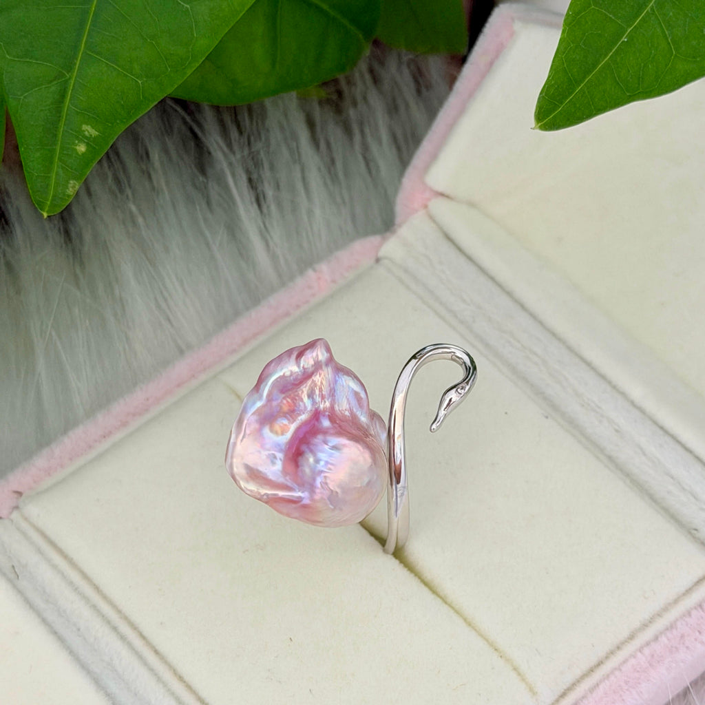 A silver ring shaped like a swan, featuring a large, lustrous pink baroque pearl on its beak, displayed inside an open cream-colored jewelry box with green leaves in the background.