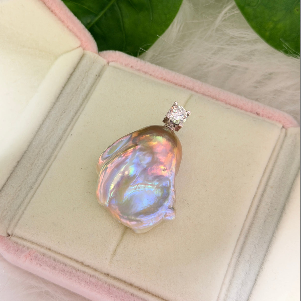 A baroque pearl pendant with iridescent rainbow sheen, set in a silver-toned prong with a clear gemstone, displayed inside an open pink velvet jewelry box.