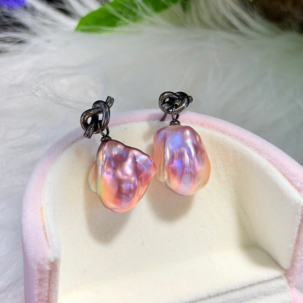 A pair of baroque pearl earrings with iridescent pink, purple, and blue pearl, suspended from dark metal posts, displayed on a pink velvet ring box.
