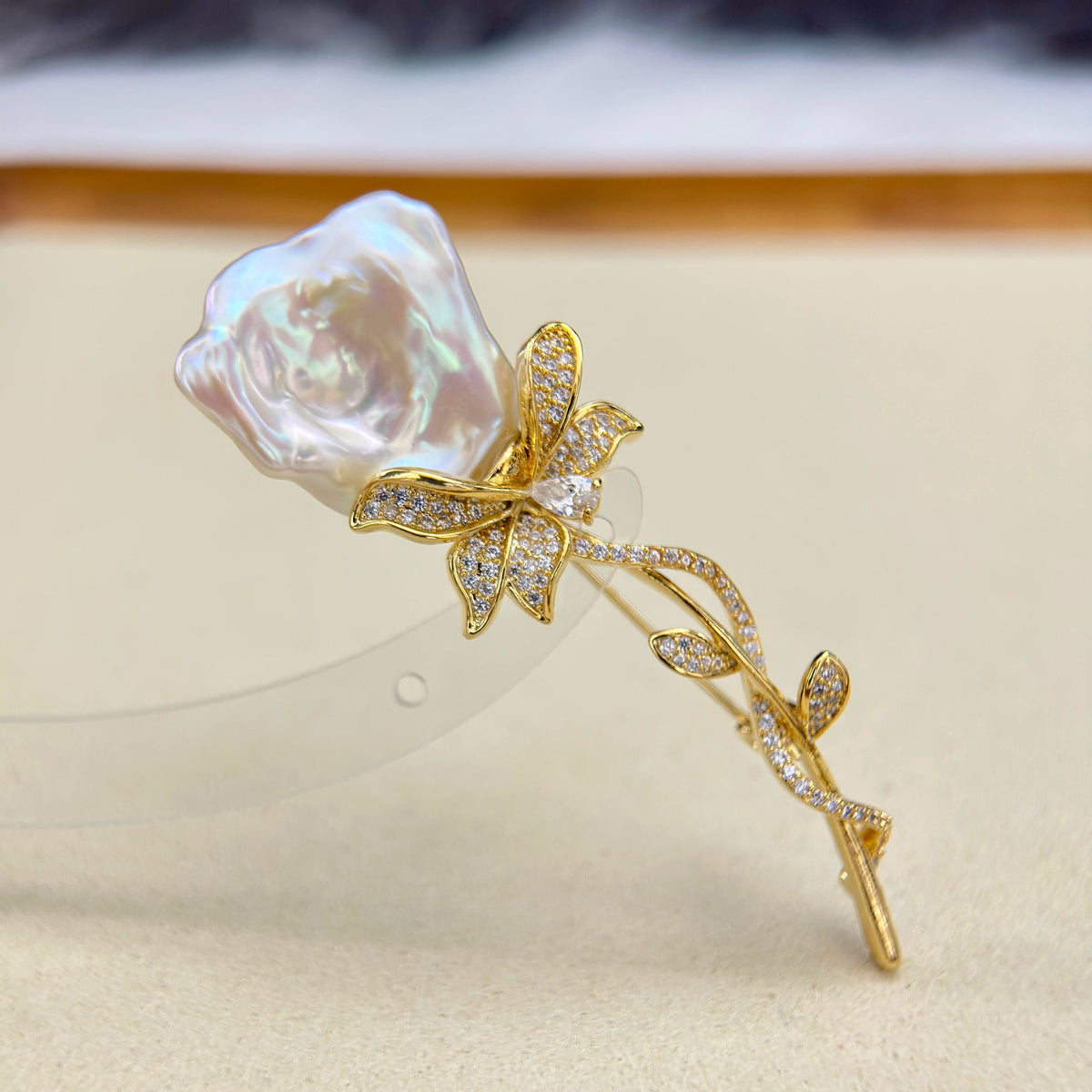 A gold rose-shaped hair claw clip featuring a large, lustrous white baroque pearl rose head and sparkling clear crystal-encrusted leaves and stem, displayed on a beige surface.