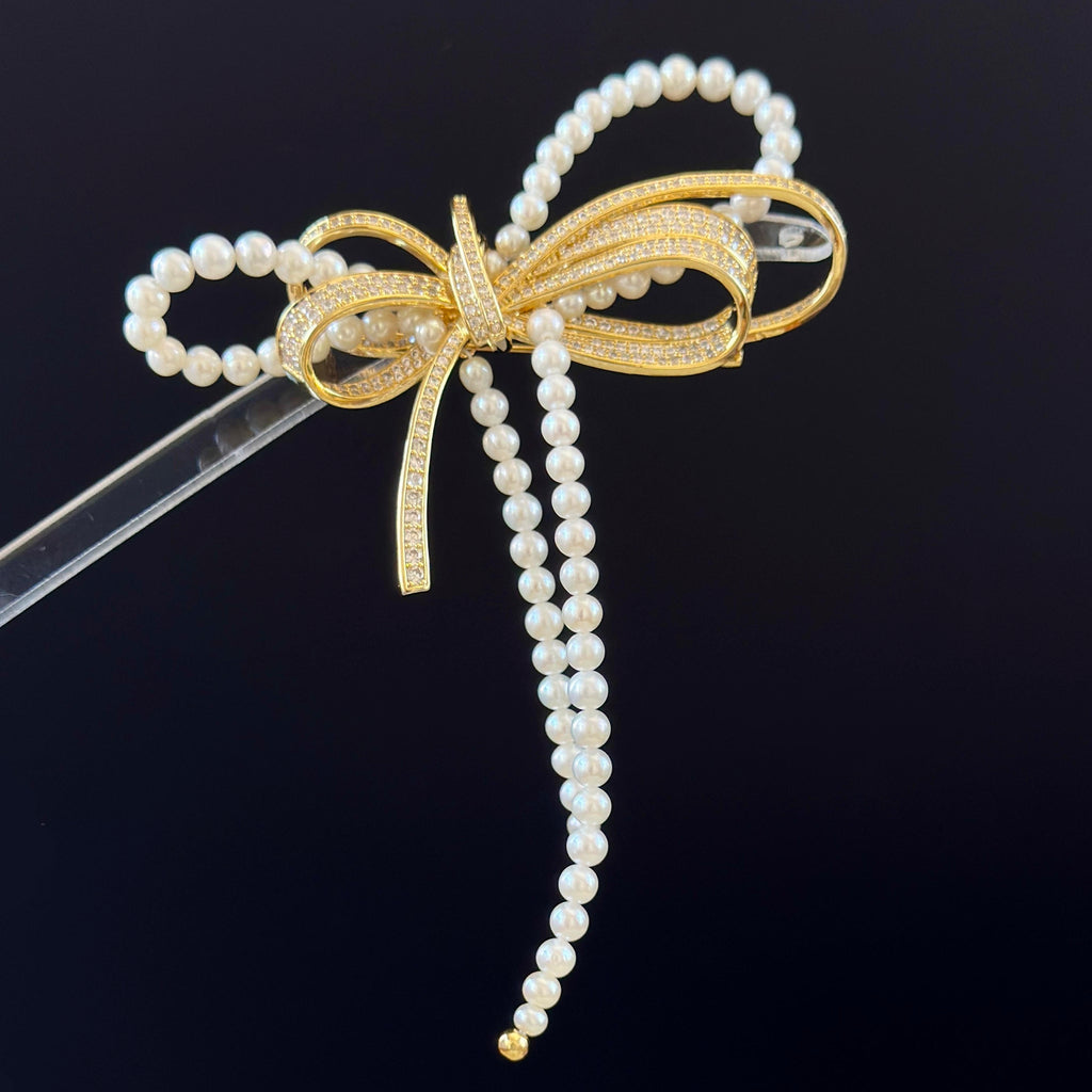 A pearl and gold bow hair accessory with dangling pearl strands, displayed on a clear stand against a dark background.